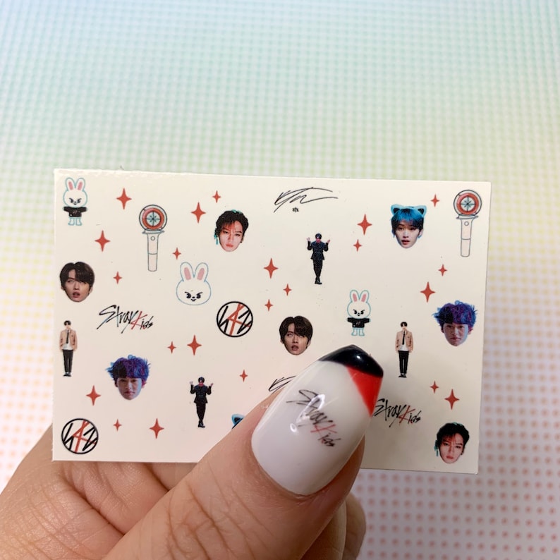 Op de afbeelding: Een vel nagelstickers met een witte achtergrond en verschillende kleurrijke ontwerpen. De ontwerpen omvatten een cartoonkonijn, een microfoon, een ster en een groep mannen. De tekst "Stray Kids" is op het vel gedrukt.