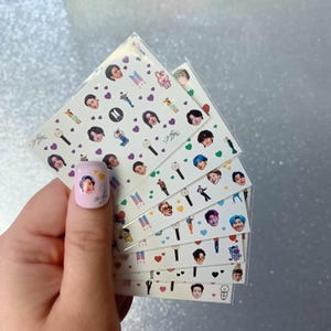以下が含まれることがあります： 音楽グループの様々な画像が特徴のネイルアートステッカーシート。ステッカーには肖像画、ハート、その他のグラフィックが含まれています。1つの爪には肖像画とサインが描かれています。ステッカーは透明なプラスチックパッケージに入っています。