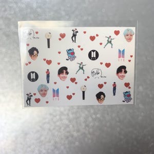 May include: A sheet of stickers featuring images of people, hearts, and logos. The stickers include faces, figures, and the BTS logo. The stickers are in a clear plastic sleeve.