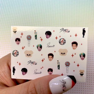 Op de afbeelding: Een vel nagelstickers met daarop de K-popband Stray Kids. De stickers bevatten afbeeldingen van de bandleden, hun logo en een cartoonhond. De stickers zijn op een witte achtergrond met rode sterren.