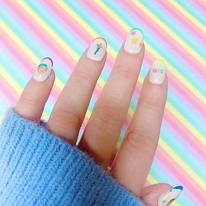 May include: A hand with painted nails featuring various designs. The nails are white with colorful accents and small images, including a person's face, a figure, and the letters "BTS". The background is a pastel rainbow pattern.