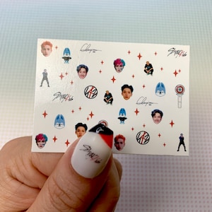 Op de afbeelding: Een vel nagelstickers met afbeeldingen van een K-pop-groep, Stray Kids. De stickers bevatten het logo van de groep, portretten van individuele leden en een cartoonwolf. Het vel bevat ook de handtekening van de groep en het woord "Stray Kids".