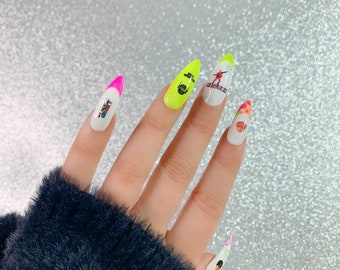May include: A hand with long, pointed artificial nails. The nails feature a variety of designs, including neon green, white, and pink tips. Some nails have small graphic images. The nails are set against a sparkly, silver background.