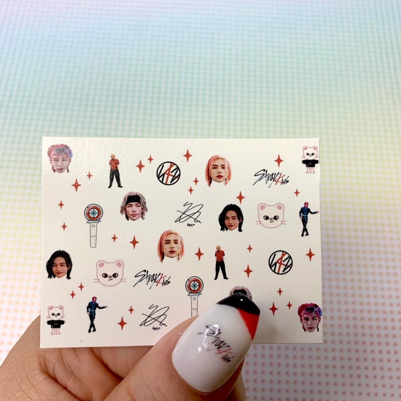 Op de afbeelding: Een vel stickers met afbeeldingen van een K-pop-groep, Stray Kids. De stickers bevatten afbeeldingen van de bandleden, het bandlogo en een cartoonkat. De stickers zijn op een witte achtergrond met rode sterren.