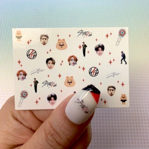 Op de afbeelding: Een vel nagelstickers met een witte achtergrond en verschillende afbeeldingen, waaronder cartoondieren, sterren en de tekst "Stray Kids".