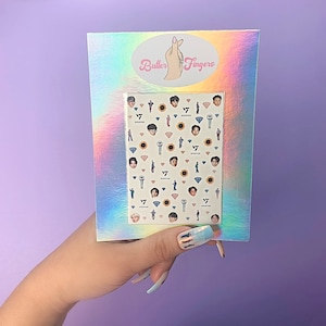 Puede incluir: Una hoja de pegatinas para uñas con un grupo de K-pop, con imágenes de los miembros del grupo, girasoles, micrófonos y diamantes. La hoja está empaquetada en una funda de plástico transparente con un fondo holográfico y el texto "Butter Fingers" impreso en la parte delantera.