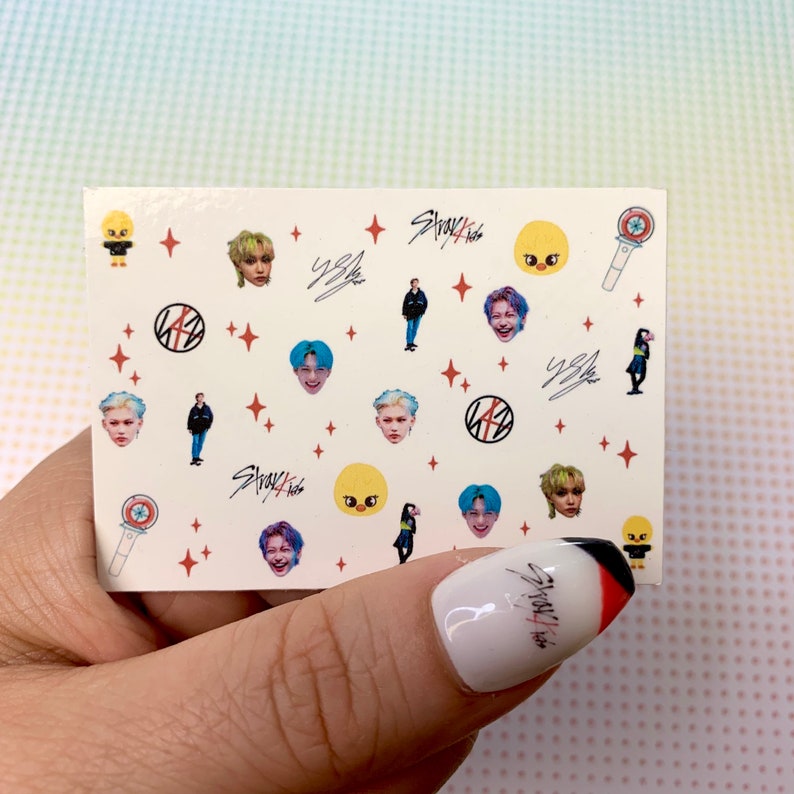 Op de afbeelding: Een vel nagelstickers met verschillende afbeeldingen, waaronder cartoonfiguren, bandlogo's en bandledenportretten. De stickers zijn op een witte achtergrond met rode sterren en de tekst "Stray Kids" in zwart.