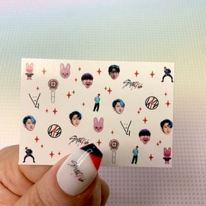 Op de afbeelding: Een vel met witte stickers met verschillende afbeeldingen, waaronder cartoonkonijnen, sterren en de gezichten van leden van de K-pop-groep Stray Kids. De stickers bevatten ook het logo van de groep en de tekst "Stray Kids".