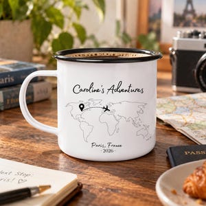 May include: A white enamel mug with a black rim featuring the text Caroline's Adventures, a black line-art world map with a dotted flight path from a location pin to an airplane icon, and the text Paris, France 2026 printed in black ink.