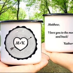 Peut inclure: Deux mugs en émail blanc avec des bords noirs sont présentés. L'un présente un cœur avec des initiales et une flèche, l'autre un message: "I love you to the moon and back!" et des noms. Un cadeau romantique.
