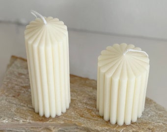 May include: Two white pillar candles with a fluted design. The candles are on a natural stone surface.