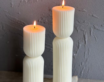 May include: Two white pillar candles with fluted designs are lit and placed on a stone surface. The candles are different sizes and have a warm glow.