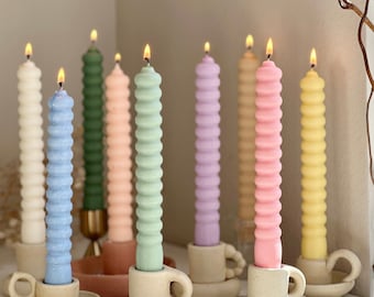 May include: Nine pastel colored candles with a wavy texture are lit and placed in ceramic cup-shaped candle holders. The holders are beige and have a saucer base.