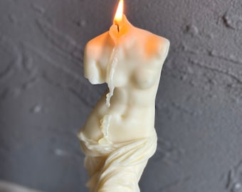 Puede incluir: Una vela blanca con forma de estatua de mujer, con una llama que arde en la parte superior. La vela está sobre una superficie marrón claro.