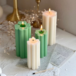 May include: Four candles with a flower-shaped top. Two candles are green and two are white. The candles are on a white surface with a piece of paper underneath.