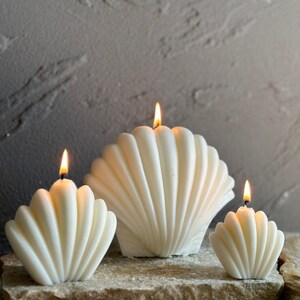 May include: Three white shell-shaped candles, each with a lit wick. The candles are arranged in a row from largest to smallest.