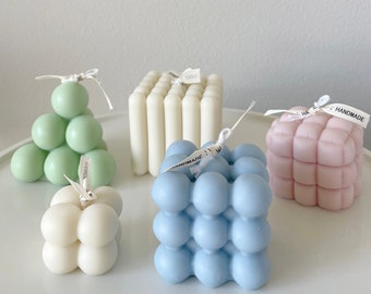 Puede incluir: Cinco velas artesanales de diferentes formas y colores. Las velas tienen forma de pirámide, cubo y esfera. Las velas son verdes, blancas, azules y rosas.