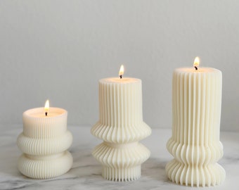 May include: Three white pillar candles with a fluted design. The candles are lit and are on a white marble surface.