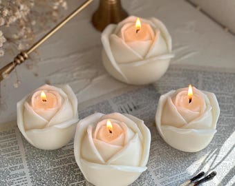 Può includere: Quattro candele bianche a forma di rosa, ciascuna con un solo stoppino. Le candele sono accese e le fiamme sono visibili. Le candele sono su una superficie bianca con un giornale sotto.