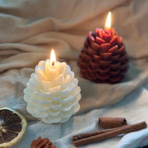 Może przedstawiać: Dwa zapalone świece w kształcie szyszki, jedna biała i jedna brązowa, leżą na neutralnej kolorystycznie tkaninie. Obecny jest również plaster suszonej cytryny, mała szyszka i dwa patyczki cynamonu. Świece są przedmiotem dekoracyjnym.