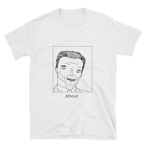 May include: A white t-shirt featuring a black and white line drawing of Arnold Schwarzenegger's face within a square frame. The text "ARNOLD" is positioned beneath the frame.
