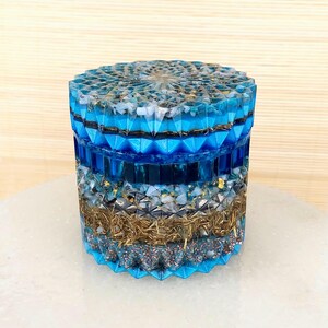 Könnte beinhalten: Eine runde, mehrschichtige, blaue und goldene Harz-Schmuckschatulle mit geometrischem Design. Die Schachtel ist mit verschiedenen Kristallen und Goldflocken gefüllt.