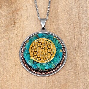 Könnte beinhalten: Halskette mit silberfarbener Kette und rundem Anhänger. Der Anhänger zeigt ein goldfarbenes Flower-of-Life-Design in der Mitte, umgeben von türkisfarbenen Steinen und einem kupferfarbenen Perlenrand. Der Hintergrund ist eine hellbraune Holzoberfläche.
