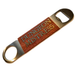 Könnte beinhalten: Ein brauner Flaschenöffner aus Holz mit einem Metalldeckel. Der Griff ist mit den Worten "Dungeon Mistress" in goldenen Buchstaben graviert.