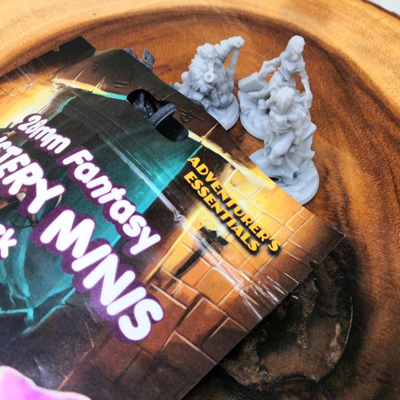 Pode incluir: Um close-up de um pacote de Miniaturas de Fantasia de 28 mm da Adventurer's Essentials. O pacote est&aacute; aberto e mostra tr&ecirc;s figuras em miniatura de pl&aacute;stico cinza de um an&atilde;o, uma guerreira e uma elfa.