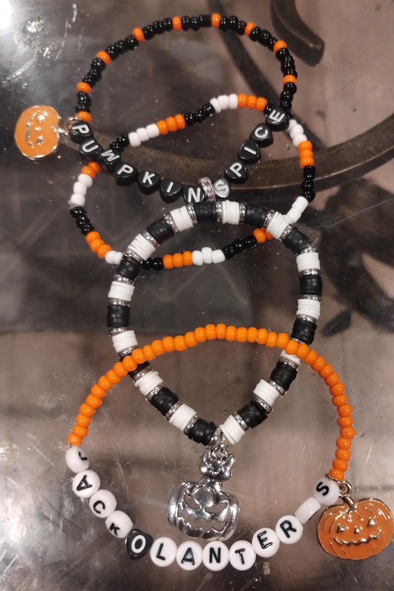 Op de afbeelding: Drie kralenarmbanden met pompoenbedels. De armbanden zijn van zwarte, witte en oranje kralen. De eerste armband spelt "Pumpkin Spice", de tweede armband spelt "Jack O Lanterns" en de derde armband heeft zwarte en witte kralen.