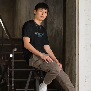 May include: A man wearing a black t-shirt with the text "Wikipedia Editor" in blue lettering. He is sitting on a stool in a room with a concrete wall.