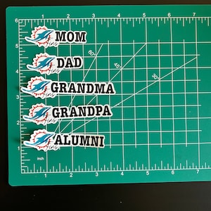 May include: Five stickers with a dolphin logo and the words "MOM Fins", "DAD Fins", "GRANDMA Fins", "GRANDPA Fins", and "ALUMNI Fins". The stickers are on a green cutting mat with inch markings.
