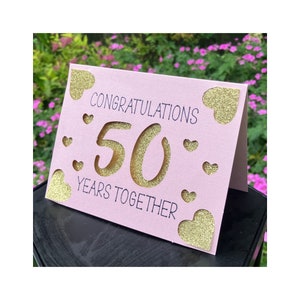 Puede incluir: Tarjeta rosa con corazones de purpurina dorada y el texto "Felicidades 50 a&ntilde;os juntos".