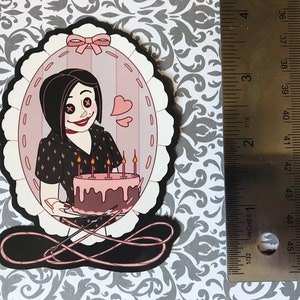 May include: A die-cut sticker featuring a cartoon character holding a cake with lit candles. The character is inside a pink oval frame with a black border. The sticker is approximately 3 inches tall.