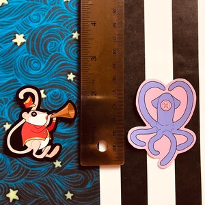 May include: Two stickers: a mouse in a red uniform playing a horn and a pink and purple squid with a heart-shaped head. A ruler shows the stickers are approximately 2 inches in height. The background is a mix of blue, black, and white.