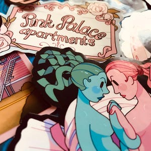 May include: A collection of colorful stickers featuring various designs. One sticker reads "Pink Palace apartments" in a decorative font. Other stickers depict illustrations of figures and buildings, with a focus on a couple holding hands.