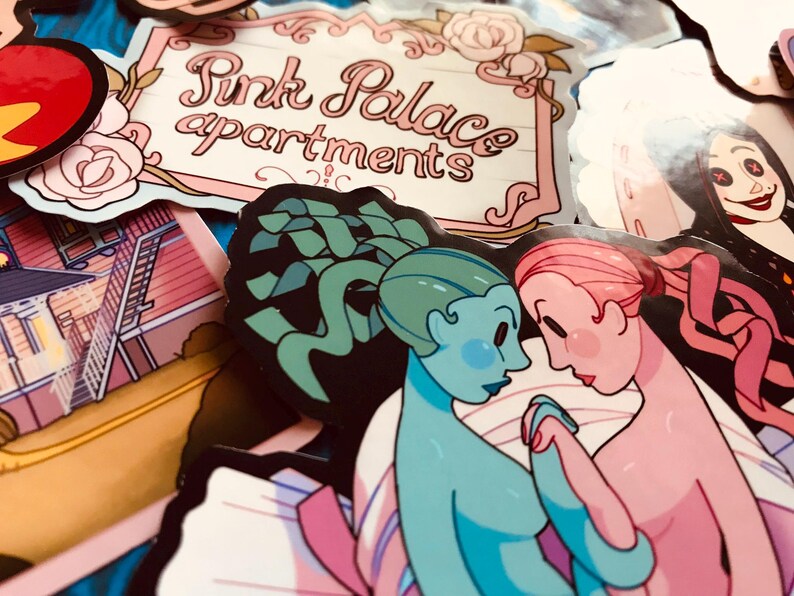 May include: A collection of colorful stickers featuring various designs. One sticker reads "Pink Palace apartments" in a decorative font. Other stickers depict illustrations of figures and buildings, with a focus on a couple holding hands.