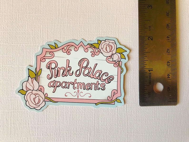 May include: A pastel blue and pink sign with a white wooden background and gold trim. The sign reads "Pink Palace apartments" with a floral design around the edges.