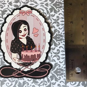 May include: A die-cut sticker featuring a cartoon character holding a cake with lit candles. The character is inside a pink oval frame with a bow at the top. The sticker is approximately 3 inches tall.