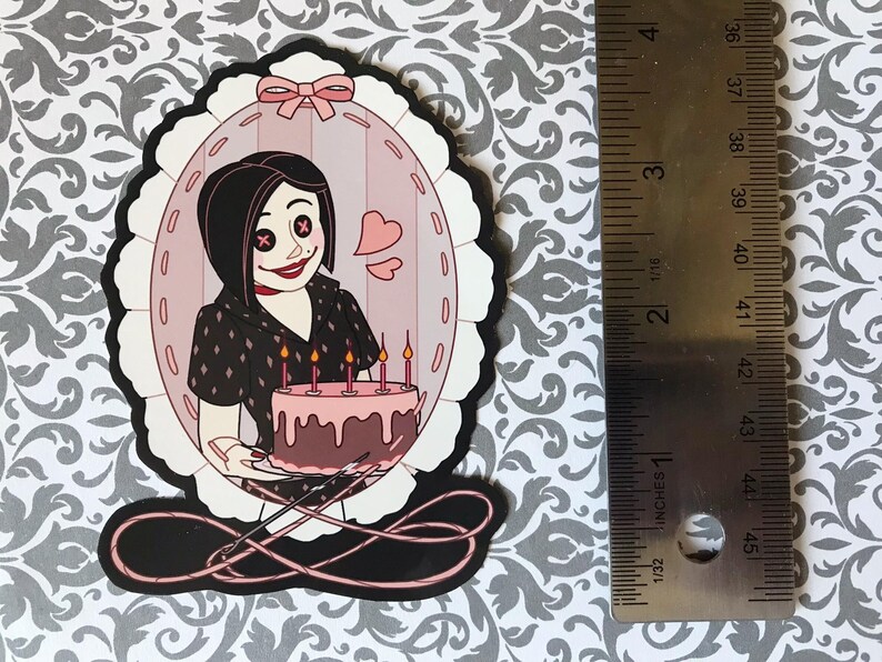 May include: A die-cut sticker featuring a cartoon character holding a cake with lit candles. The character is inside a pink oval frame with a bow at the top. The sticker is approximately 3 inches tall.