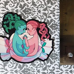 May include: A sticker featuring two cartoon figures, one green and one pink, embracing inside a candy wrapper. The green figure has a green and blue hair, and the pink figure has pink hair. The candy wrapper is white with pink, blue, and green stripes.