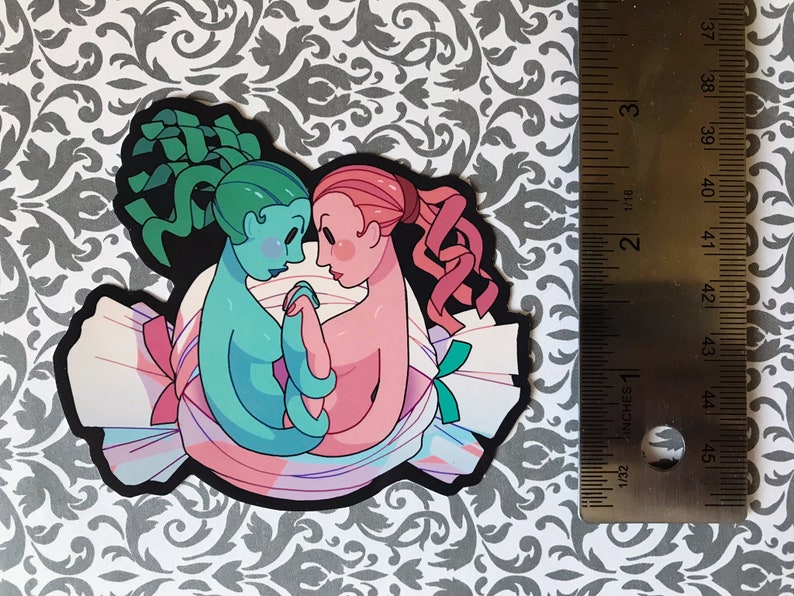 May include: A sticker featuring two cartoon figures, one green and one pink, embracing inside a candy wrapper. The green figure has a green and blue hair, and the pink figure has pink hair. The candy wrapper is white with pink, blue, and green stripes.