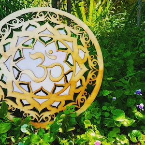 Puede incluir: Un mandala de madera con un símbolo Om tallado en el centro. El mandala está rodeado por un borde decorativo y se coloca sobre un fondo de follaje verde.