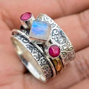 Puede incluir: Un anillo de plata con una piedra de luna y dos piedras preciosas rojas. El anillo tiene un diseño decorativo y en espiral.
