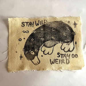 Könnte beinhalten: Ein beigefarbener Stoff-Patch mit einer Schwarz-Weiß-Illustration eines Schnabeltiers. Der Text "Stay Wild" und "Stay Weird" ist auf dem Patch aufgedruckt.