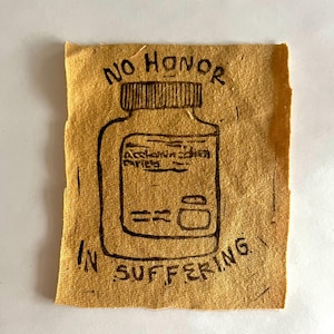 Puede incluir: Un parche de tela marrón con tinta negra que representa una botella de píldoras con el texto "NO HONOR IN SUFFERING" y las palabras "ACHILLES'S HEEL" en la etiqueta.
