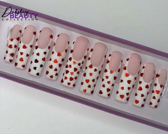 May include: A set of artificial nails with a white base, red heart-shaped accents, and a pink tip. The nails are arranged in a rectangular box. The brand name "Dakshay Beauty Collection" is visible in the upper left corner.