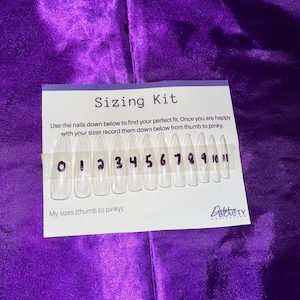 May include: Sizing kit for press-on nails with numbered clear plastic nail tips. The kit includes 11 sizes, numbered 0 through 10. The text on the card reads "Sizing Kit" and "Use the nails down below to find your perfect fit. Once you are happy with your sizes record them down below from thumb to pinky. My sizes (thumb to pinky):"