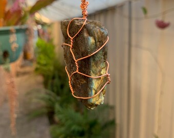 May include: A copper wire wrapped labradorite pendant on a brown cord necklace. The labradorite is a dark grey with iridescent blue and green flashes.