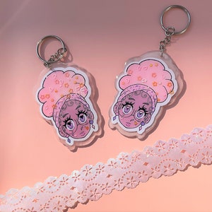 May include: Two pink and white keychains with a cartoon illustration of a person with pink hair and a pink and white patterned headband. The keychains are made of clear plastic and have a sparkly finish.